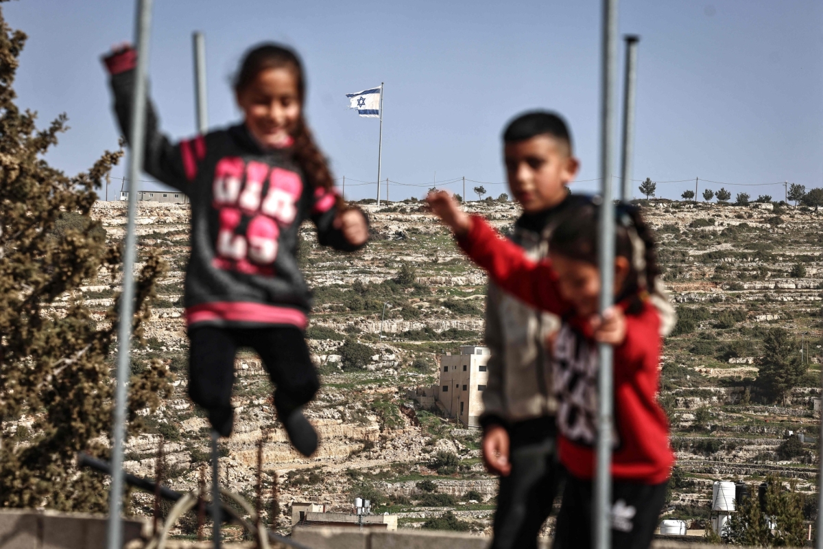 Palestinian children play on a trampoline in Umm Safa village, north of Ramallah in the occupied West Bank, opposite an Israeli flag that was raised on a hilltop overlooking the village on 16 February 2026.