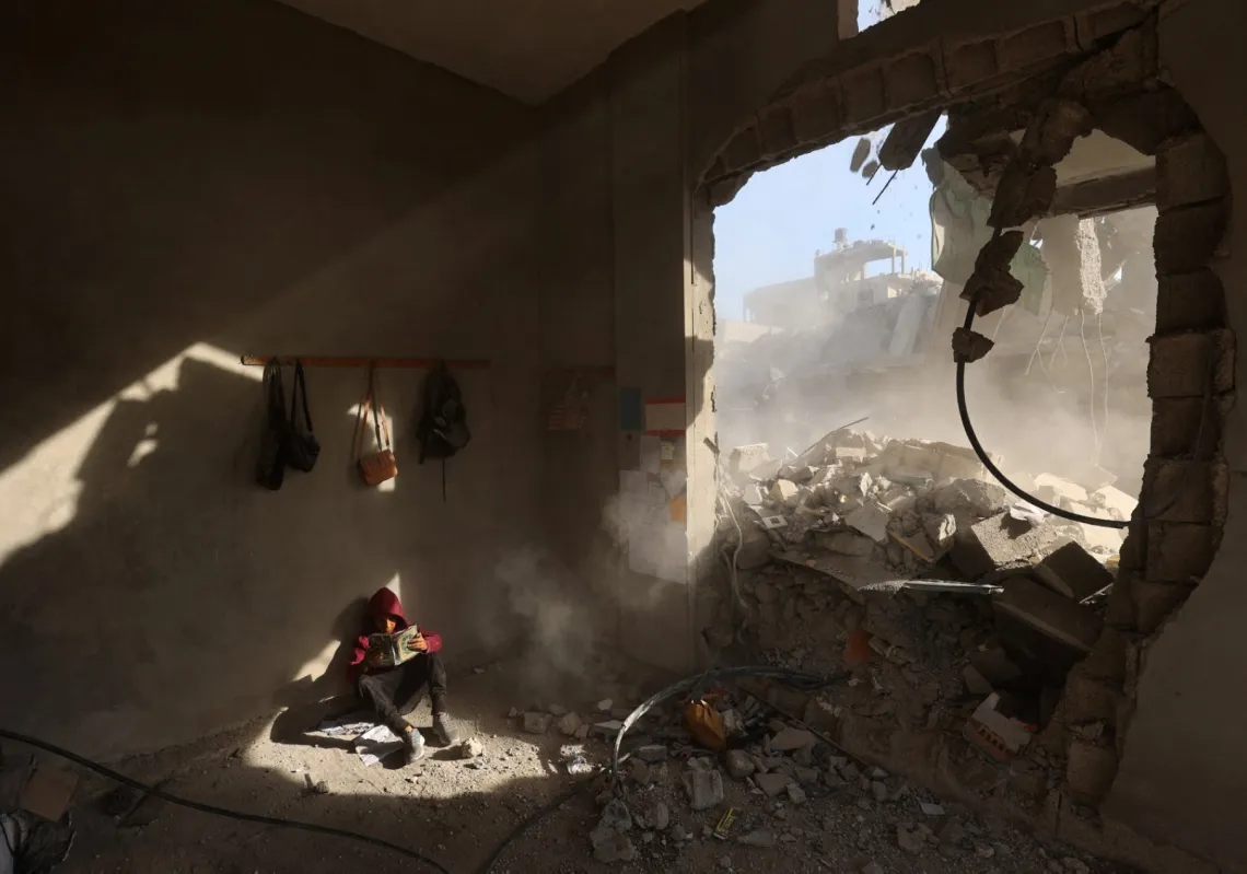 طفل فلسطيني يجلس بين أنقاض منزل ممسكا بكتاب بعد غارة ليلية على مخيم النصيرات وسط غزة، 29 أبريل 2025