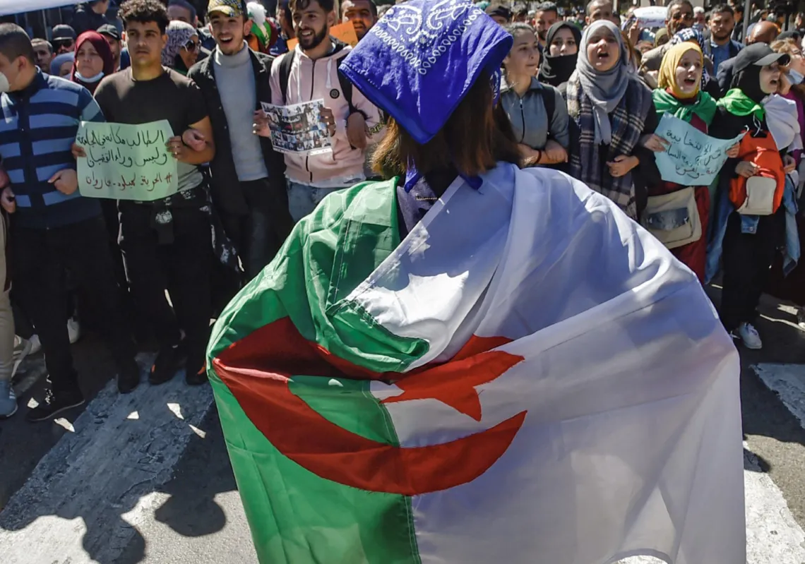 امرأة ملتفة بالعلم الوطني الجزائري تقف أمام طلاب يُشاركون في مظاهرة بالعاصمة الجزائرية، الجزائر، في 16 مارس2021