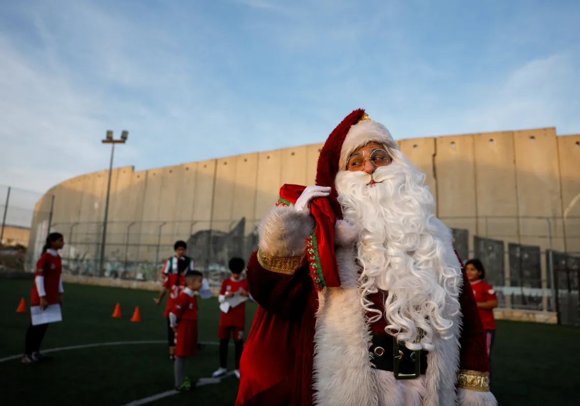 A man dressed as Santa reacts next to Palestinian teens playing next to the Israeli apartheid wall separating the Aida refugee camp from Jerusalem, in the Israeli-occupied city of Bethlehem, on 22 December 2025.