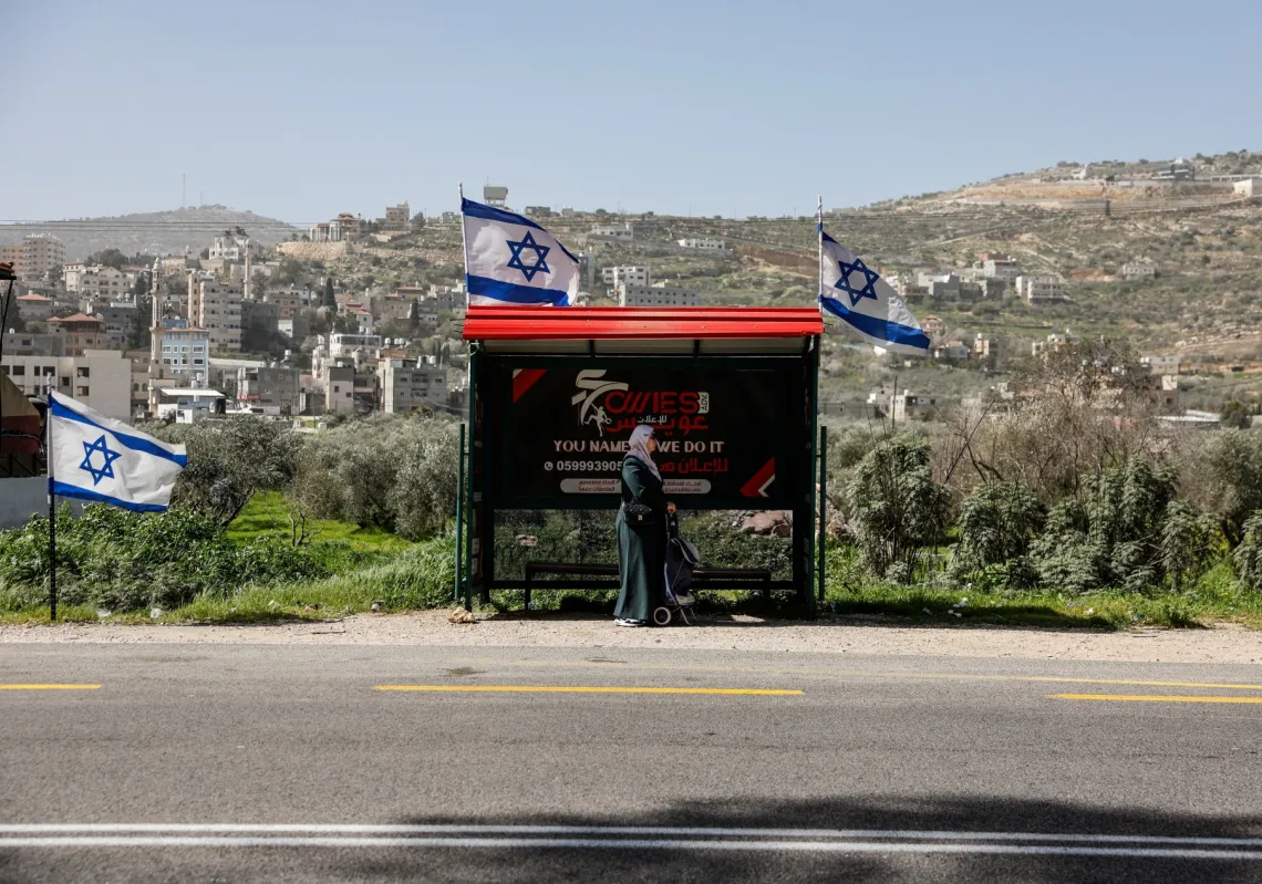 امرأة فلسطينية تنتظر عند موقف حافلات بجانب أعلام إسرائيلية، قرب بلدة اللبن الشرقية بين نابلس ورام الله
