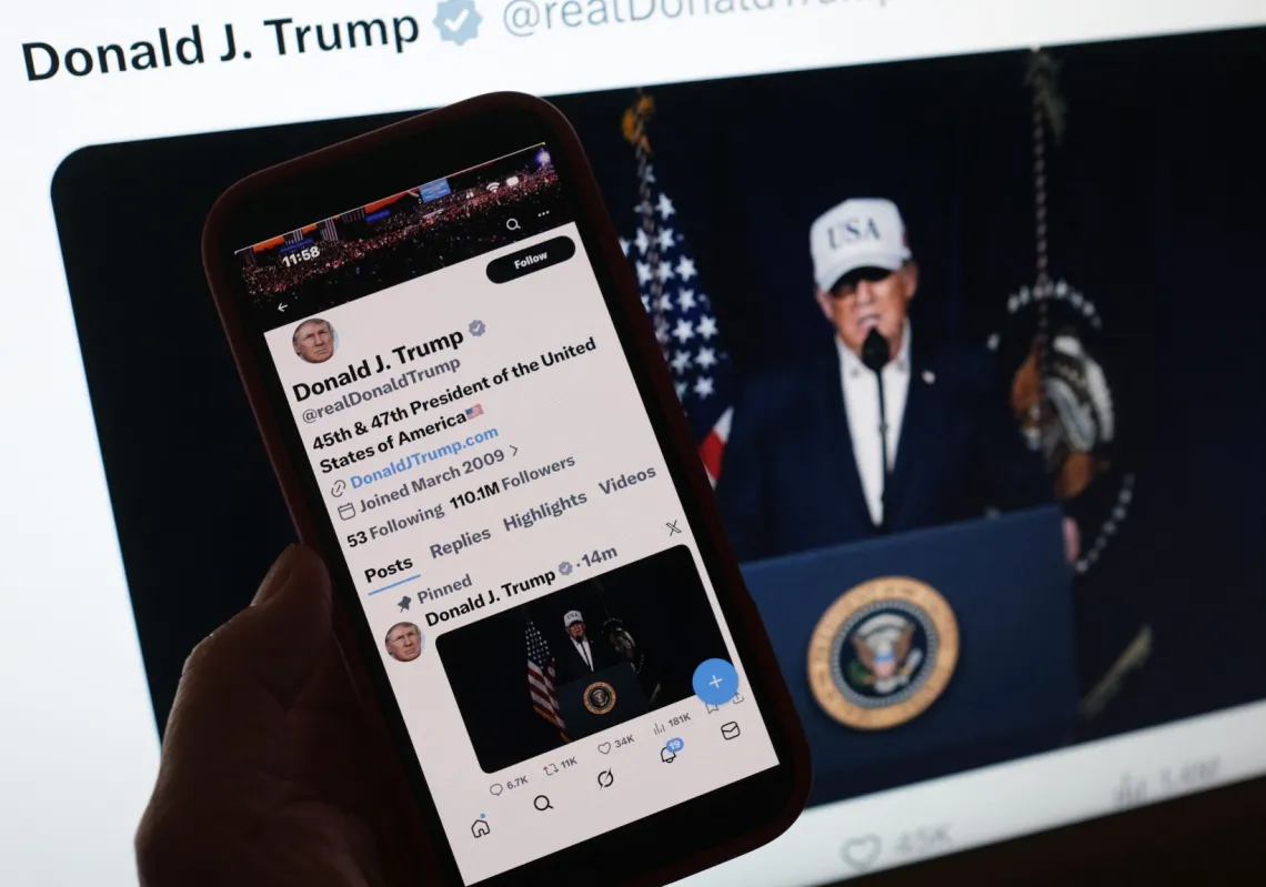 شخص ينظر إلى هاتف ذكي يعرض بيان الرئيس الأميركي دونالد ترمب بدء العمليات القتالية في إيران، إلى جانب شاشة كمبيوتر تعرض الرسالة نفسها المنشورة على موقع X بتاريخ 28 فبراير/شباط 2026. 