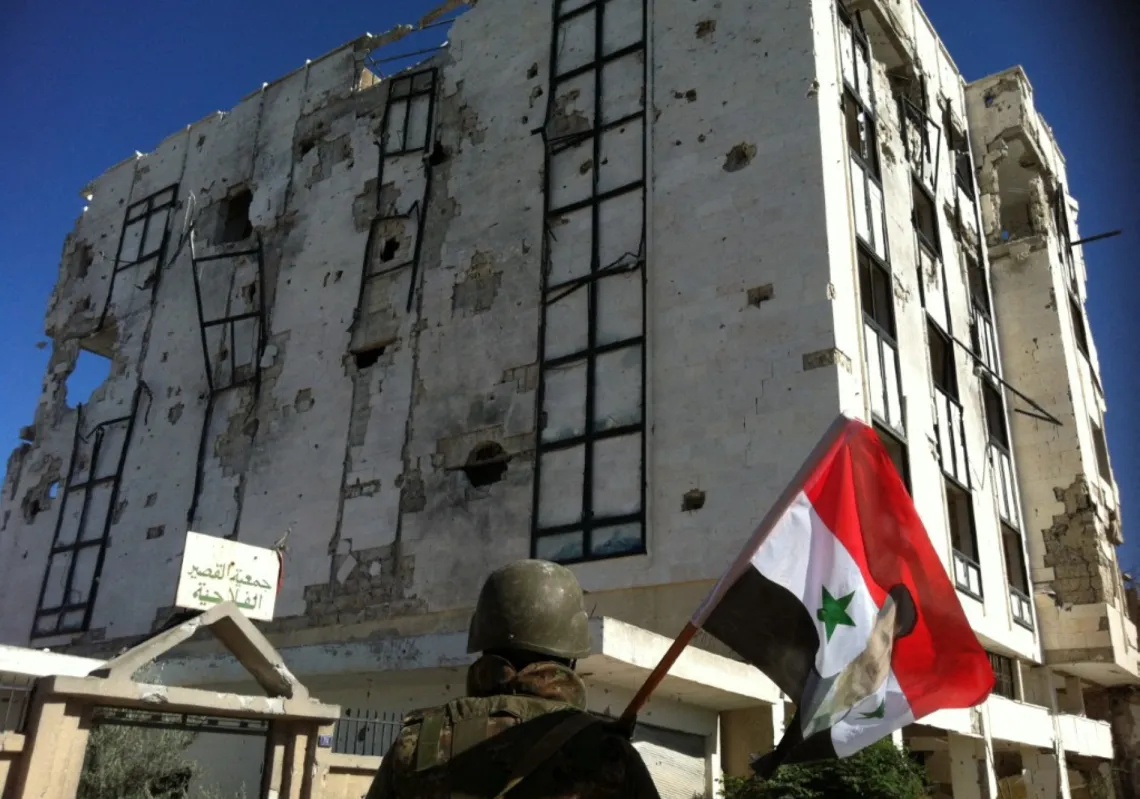 A Syrian army soldier holds a national flag featuring Syria's President Bashar al-Assad in front of a building left in ruins on 5 June 2013 in the city of Qusayr in the Homs province. 