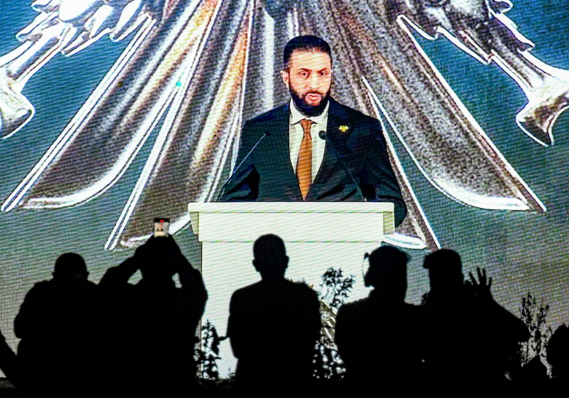 أشخاص يتابعون خطاب الرئيس السوري أحمد الشرع على شاشة عملاقة قرب ضريح الجندي المجهول في جبل قاسيون المطل على دمشق، خلال مراسم الكشف عن الشعار الوطني الجديد، 3 يوليو 2025