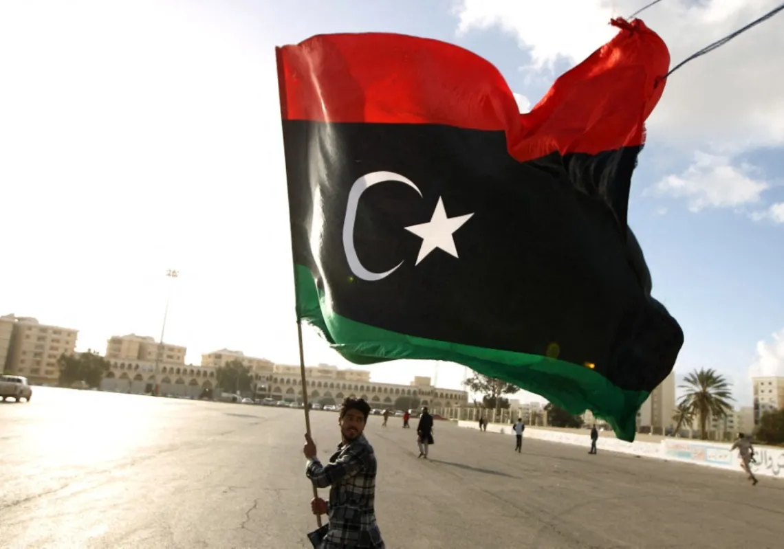 رجل ليبي يلوّح بعلم بلاده بينما يتجمّع متظاهرون لمطالبة المجتمع الدولي برفع الحظر المفروض على تسليح الجيش الليبي أو تزويده بالذخيرة، وذلك في مدينة بنغازي الساحلية الشرقية في 27 فبراير 2015
